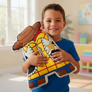 Puede incluir: Un coj&iacute;n de peluche con la forma del personaje Woody de la pel&iacute;cula Toy Story. El coj&iacute;n presenta una camisa amarilla con cuello rojo, chaleco marr&oacute;n y una placa de sheriff. El coj&iacute;n es sostenido por una persona que lleva una camiseta azul.
