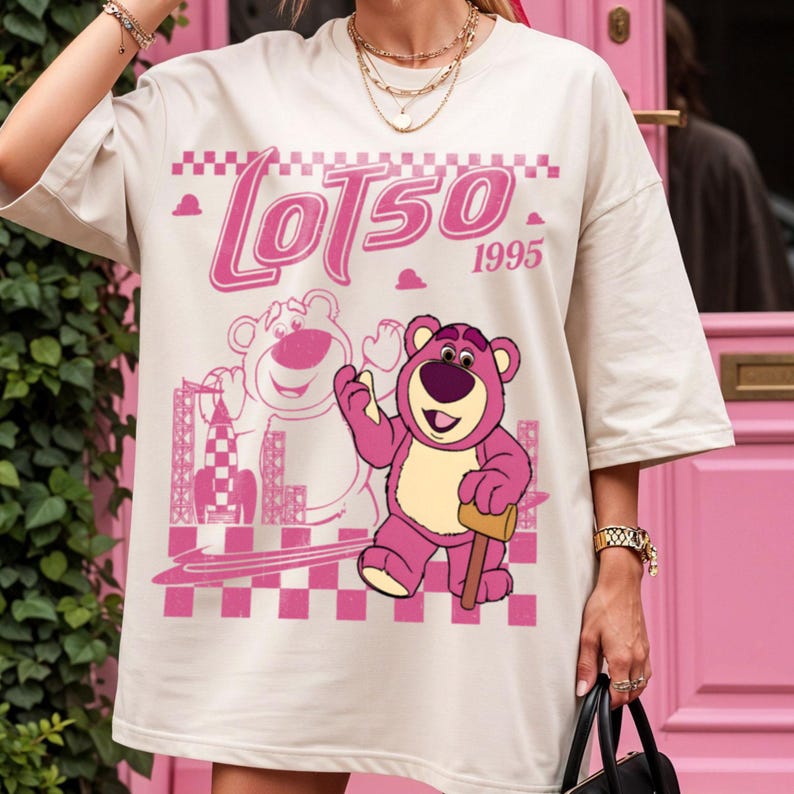 May include: Cream-colored t-shirt featuring a pink graphic of a bear character with the text "Lotso 1995". The design includes a cityscape and a checkered pattern, creating a playful and nostalgic aesthetic.