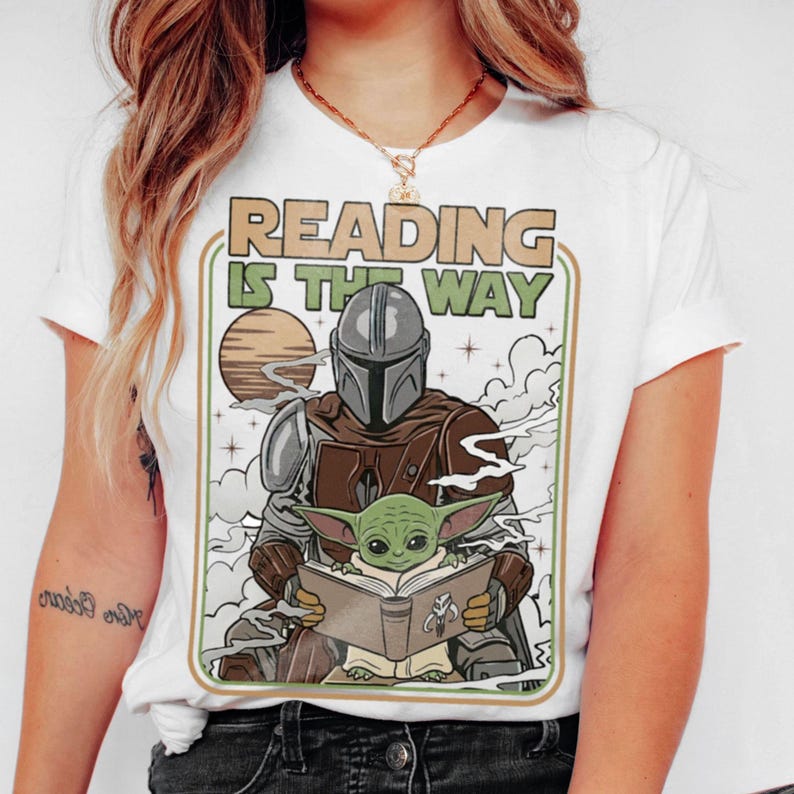 Puede incluir: Camiseta blanca con un gr&aacute;fico de The Mandalorian y Grogu leyendo un libro, con el texto "READING IS THE WAY" en un marco rectangular. El dise&ntilde;o incluye un planeta y nubes.