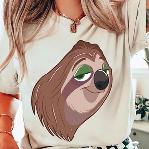 May include: Cream-colored t-shirt featuring a cartoon sloth face with brown fur, green eyes, and a black nose. The sloth has a relaxed expression. The shirt is a casual style.