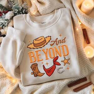 Puede incluir: Sudadera blanca con dise&ntilde;o bordado de tem&aacute;tica vaquera. Presenta un sombrero de vaquero, bota, pa&ntilde;uelo, herradura y estrella de sheriff, con las palabras "And Beyond" en naranja. La sudadera est&aacute; sobre una manta color crema.