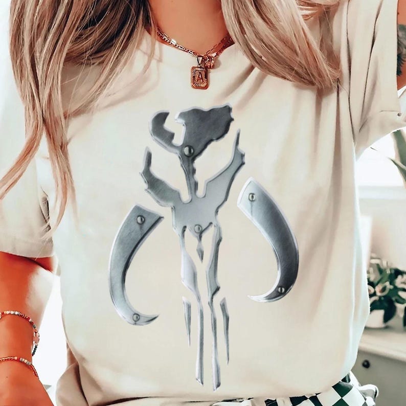 May include: A cream-coloured t-shirt with a metallic silver emblem. The emblem is a symmetrical design with curved and angular elements, resembling a helmet or mask. The t-shirt is a casual wear item.