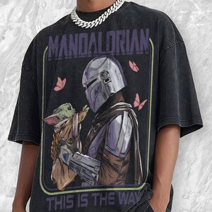 May include: Black t-shirt featuring a graphic of The Mandalorian and Grogu, with the text "MANDALORIAN" and "THIS IS THE WAY". The design includes pink butterflies. The shirt has a vintage, distressed look.