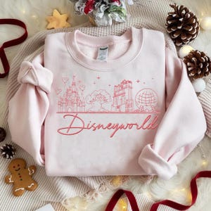 May include: A light pink sweatshirt with a red embroidered Disneyworld design. The design includes a castle, tree, tower, and globe. The word "Disneyworld" is written in cursive script below the design. The sweatshirt is folded on a cream-colored surface.