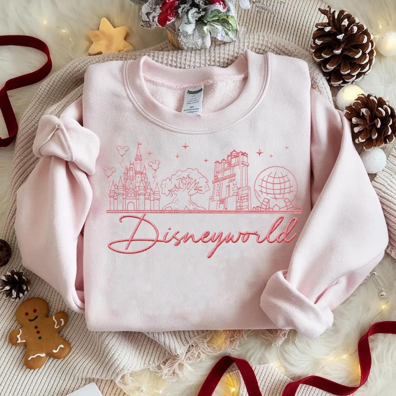 May include: A light pink sweatshirt with a red embroidered Disneyworld design. The design includes a castle, tree, tower, and globe. The word "Disneyworld" is written in cursive script below the design. The sweatshirt is folded on a cream-colored surface.