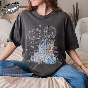 May include: Dark gray t-shirt featuring a graphic of a castle, a princess, and Mickey Mouse ears. The design includes purple and blue accents. The shirt has the words "Comfort Colors" and "Pepper" printed on it.