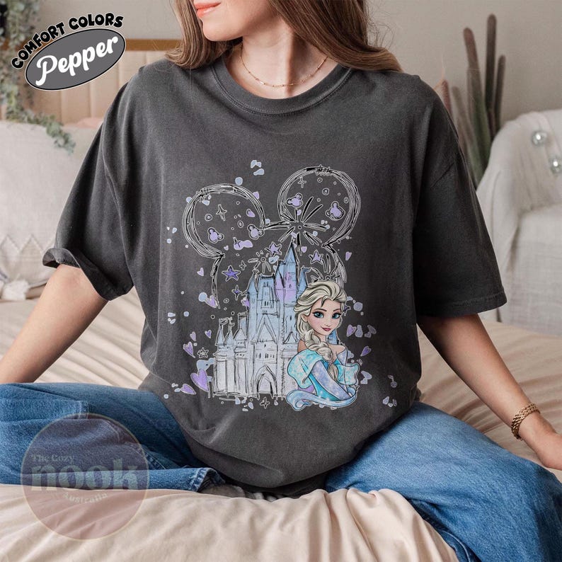 May include: Dark gray t-shirt featuring a graphic of a castle, a princess, and Mickey Mouse ears. The design includes purple and blue accents. The shirt has the words "Comfort Colors" and "Pepper" printed on it.