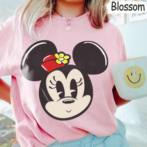 Peut inclure: Un sweat-shirt rose clair avec un grand dessin de Minnie Mouse. Le visage de Minnie est noir, avec un n&oelig;ud rouge et une fleur jaune. Le mot "Blossom" est dans le coin sup&eacute;rieur droit. Une tasse blanche avec un smiley jaune est tenue.