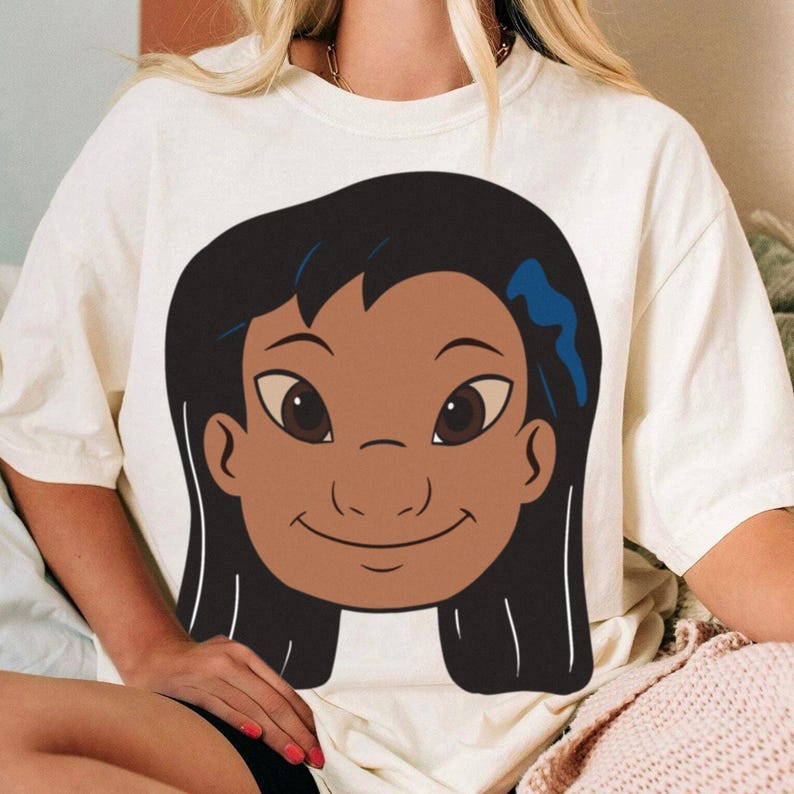Puede incluir: Camiseta de color crema con una ilustraci&oacute;n de dibujos animados de una ni&ntilde;a sonriente con cabello oscuro y ojos marrones. El gr&aacute;fico est&aacute; centrado en la camiseta, mostrando un dise&ntilde;o de personaje amigable. La camiseta es de material suave.