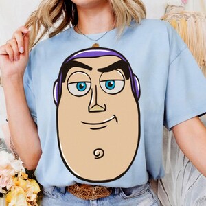 May include: Light blue t-shirt featuring a cartoon graphic of a character's face with blue eyes, black eyebrows, and a purple headband. The character's face is tan with a black outline. The t-shirt is made of a soft material.