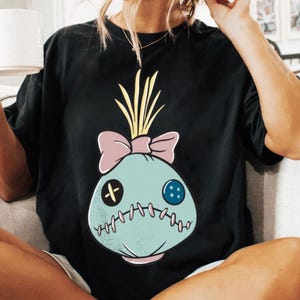 May include: Black t-shirt featuring a cartoon-style voodoo doll design. The doll is light blue with a pink bow, yellow hair, and stitched details. A button and cross are on the doll's face. The shirt is a casual, comfortable style.