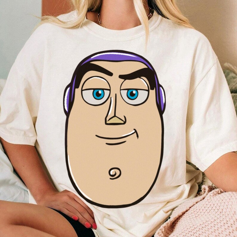 May include: Cream-colored t-shirt featuring a large cartoon graphic of Buzz Lightyear's face. The graphic includes blue eyes, a purple headpiece, and a smiling expression. The shirt is a casual style.