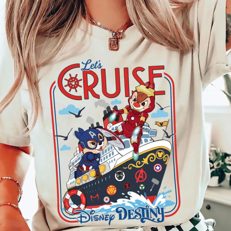 May include: Cream-colored t-shirt with a graphic featuring Chip and Dale as superheroes on a cruise ship. The text "Let's Cruise" is in red, and the ship is labeled "Disney Destiny". The design includes blue skies, birds, and waves.