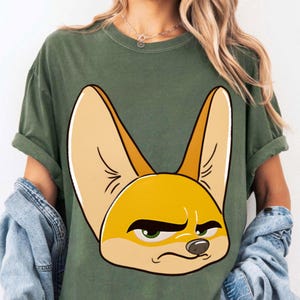 May include: Olive green t-shirt featuring a large cartoon illustration of a fennec fox's face. The fox has large, pointed ears, a yellow face, and a grumpy expression with green eyes. The shirt has short sleeves and a relaxed fit.