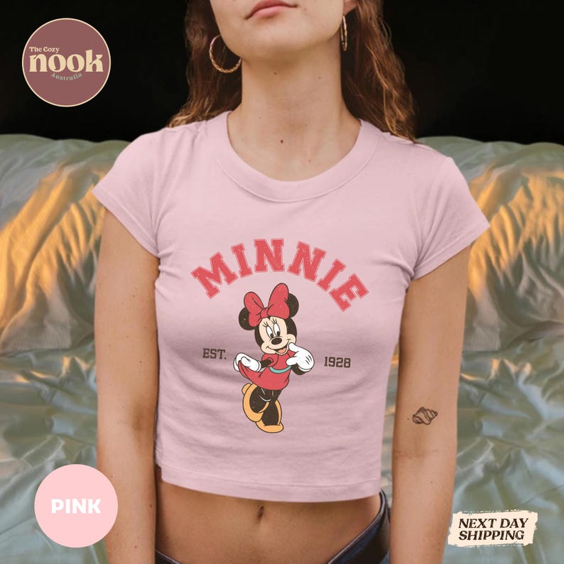 May include: Pink crop top featuring a Minnie Mouse graphic. The design includes the name "MINNIE" in red, along with an image of Minnie Mouse and the text "EST. 1928". The shirt has a crew neck and short sleeves.