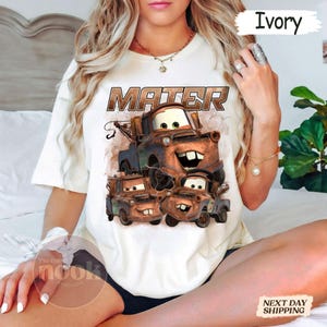 May include: Ivory-coloured t-shirt featuring a graphic of Mater from the animated film Cars. The design includes the word "MATER" in a brown, distressed font above a stack of Mater characters. The shirt is a crew neck style.