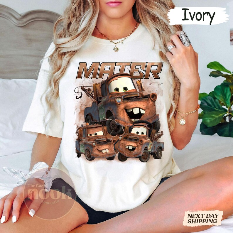 May include: Ivory-coloured t-shirt featuring a graphic of Mater from the animated film Cars. The design includes the word "MATER" in a brown, distressed font above a stack of Mater characters. The shirt is a crew neck style.