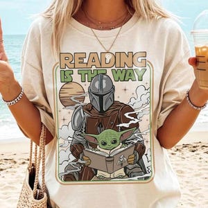 Puede incluir: Camiseta beige con un gr&aacute;fico de El Mandaloriano y Grogu leyendo un libro, con el texto "READING IS THE WAY" en un marco verde y marr&oacute;n. El dise&ntilde;o incluye una puesta de sol y nubes. La camiseta es de estilo casual.