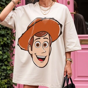 May include: Cream-colored t-shirt featuring a cartoon illustration of a smiling cowboy character with a brown hat. The graphic is centered on the front of the shirt. The shirt has short sleeves and a relaxed fit.