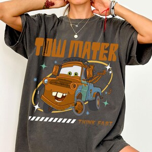 May include: A charcoal gray short-sleeve t-shirt featuring a graphic of the character Tow Mater. The design includes the text "TOW MATER" in large orange letters at the top and "THINK FAST" at the bottom, with small star accents and a yellow oval.