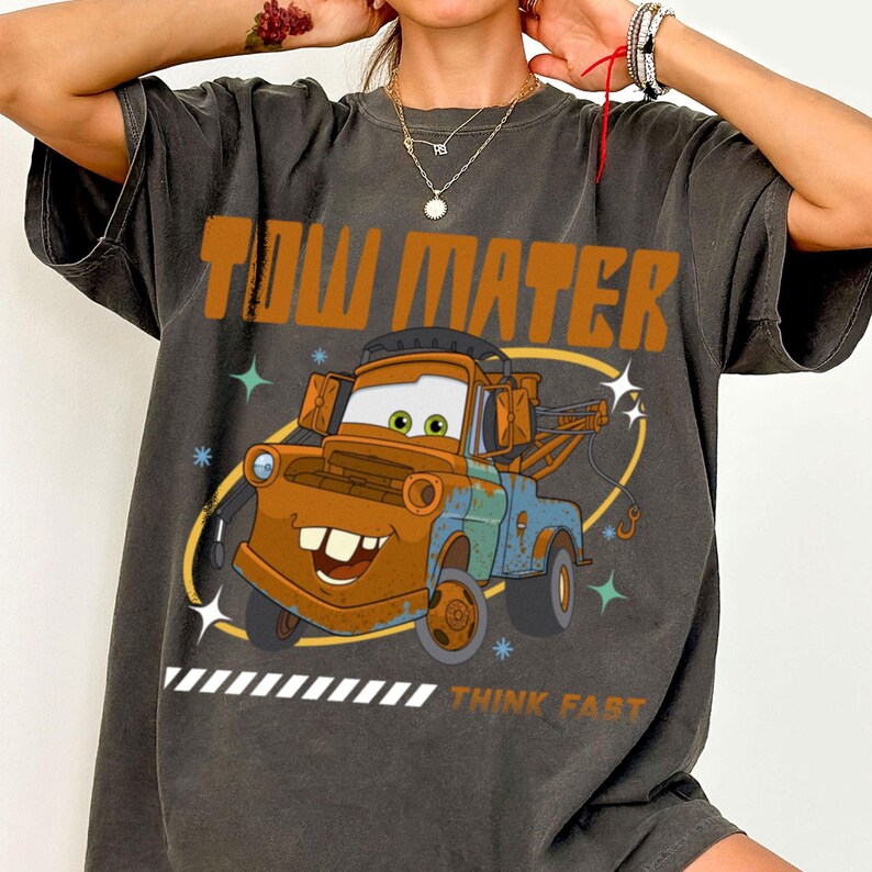 May include: A charcoal gray short-sleeve t-shirt featuring a graphic of the character Tow Mater. The design includes the text "TOW MATER" in large orange letters at the top and "THINK FAST" at the bottom, with small star accents and a yellow oval.