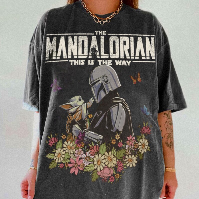 May include: Dark gray t-shirt featuring a graphic of The Mandalorian and Grogu surrounded by flowers and butterflies. The text "THE MANDALORIAN" and "THIS IS THE WAY" are printed across the top.