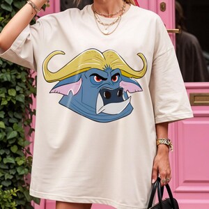 May include: Oversized beige t-shirt featuring a cartoon buffalo head with yellow horns, blue face, and red eyes. The graphic tee has a relaxed fit and short sleeves. The shirt is a casual wear item.
