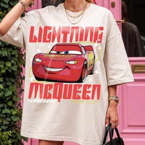 May include: A beige oversized t-shirt featuring a graphic print of the red race car Lightning McQueen. The text "LIGHTNING" appears above the car and "MCQUEEN" below it in bold red letters, with the phrase "THINK FAST" in small yellow text.
