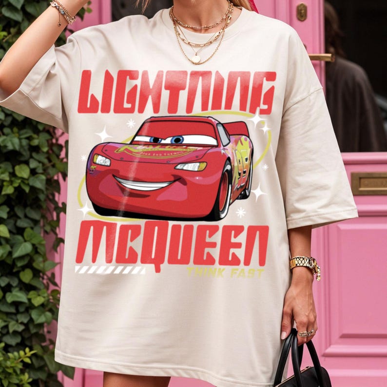 May include: A beige oversized t-shirt featuring a graphic print of the red race car Lightning McQueen. The text "LIGHTNING" appears above the car and "MCQUEEN" below it in bold red letters, with the phrase "THINK FAST" in small yellow text.
