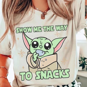 Peut inclure: T-shirt cr&egrave;me avec un personnage de dessin anim&eacute; vert tenant un petit objet. Le texte "SHOW ME THE WAY TO SNACKS" est imprim&eacute; au-dessus et en dessous du personnage en vert. Le design comprend des &eacute;toiles. V&ecirc;tement de style d&eacute;contract&eacute;.
