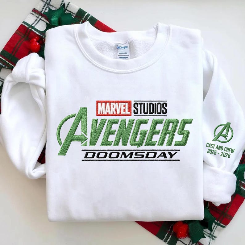 May include: White sweatshirt featuring the Marvel Studios Avengers logo in green, red, and black. The word "DOOMSDAY" is below the logo. The sleeve has the Avengers "A" logo and "CAST AND CREW 2025-2026" in green.