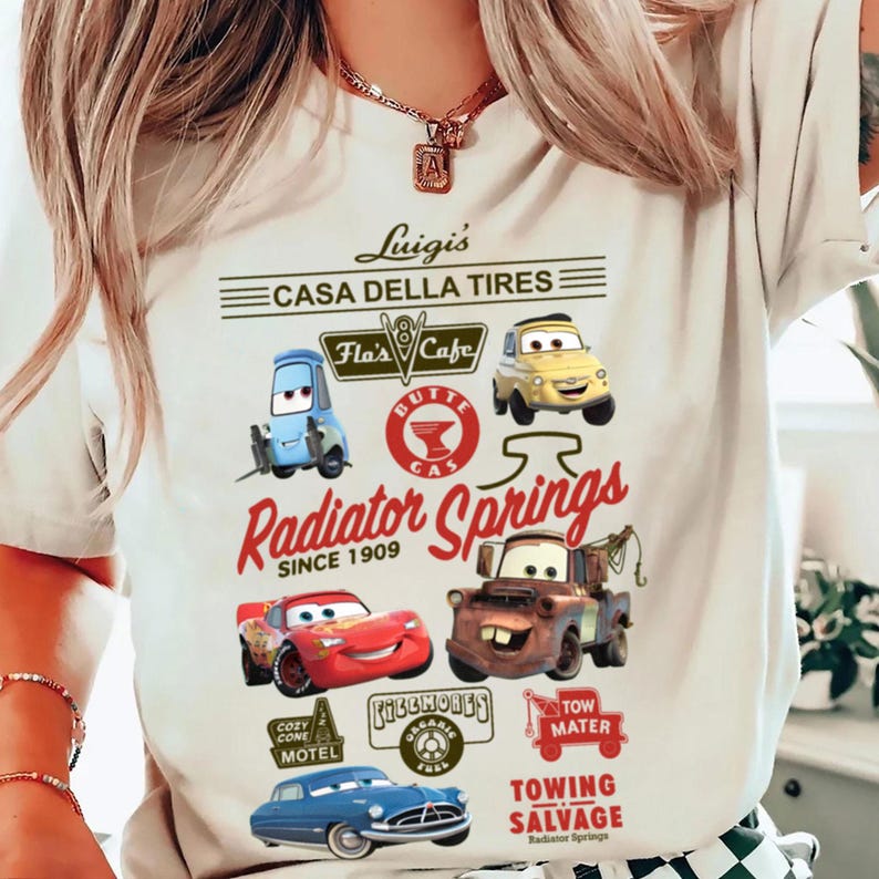 Pu&ograve; includere: Una t-shirt color crema con una stampa grafica di personaggi e luoghi del film Cars. Il design include testi come Luigi's Casa Della Tires, Radiator Springs, Flo's Cafe e Tow Mater, insieme a illustrazioni di vari veicoli.