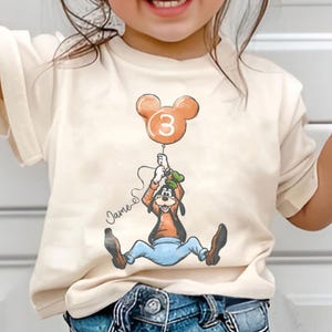 May include: Cream-colored t-shirt featuring a cartoon of Goofy holding a Mickey Mouse-shaped balloon with the number 3. The balloon is orange. The name "James" is written in cursive. The shirt is for a child.
