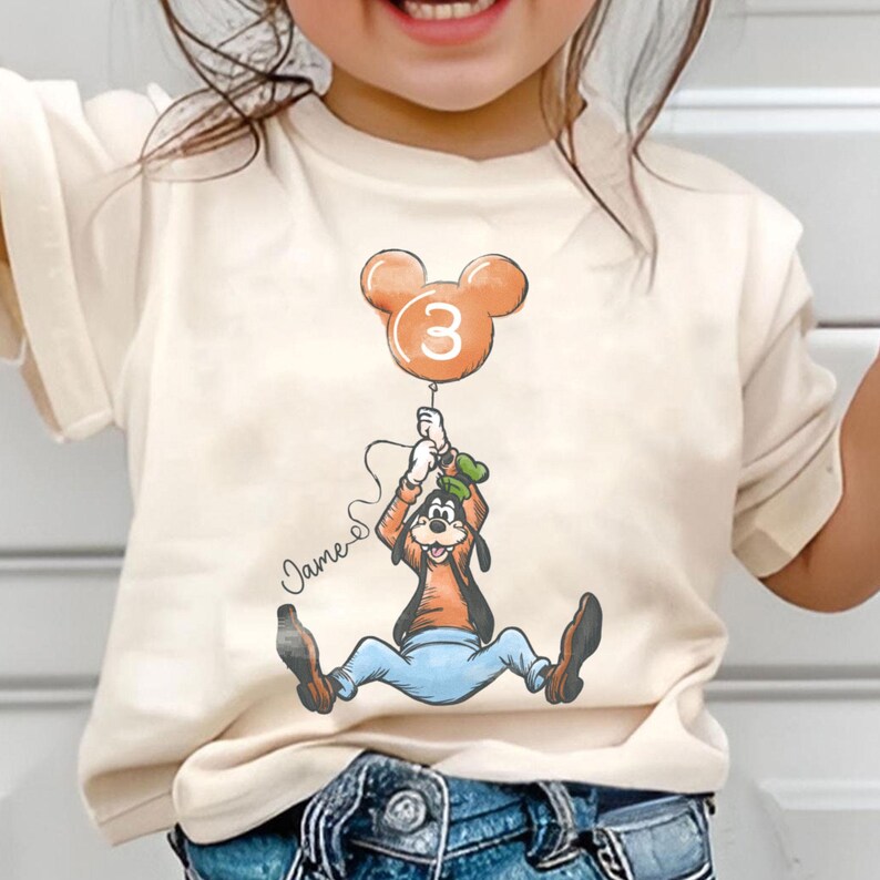 May include: Cream-colored t-shirt featuring a cartoon of Goofy holding a Mickey Mouse-shaped balloon with the number 3. The balloon is orange. The name "James" is written in cursive. The shirt is for a child.