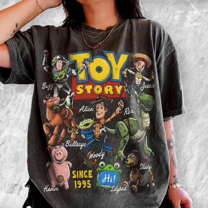 Pode incluir: Uma t-shirt cinzento-carv&atilde;o de manga curta com uma impress&atilde;o gr&aacute;fica das personagens de Toy Story, incluindo Buzz, Jessie, Bullseye, Alien, Woody, Rex, Hamm e Slinky. O texto diz Toy Story, Since 1995 e Hi, com os nomes das personagens.