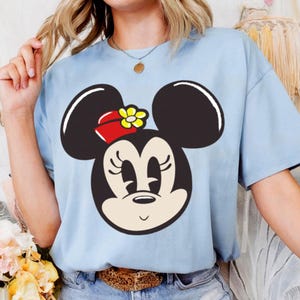 Peut inclure: T-shirt bleu clair avec un motif de Minnie Mouse. Le motif repr&eacute;sente le visage de Minnie avec des oreilles noires, un n&oelig;ud rouge et une fleur jaune. Le t-shirt est en mati&egrave;re douce et a des manches courtes.