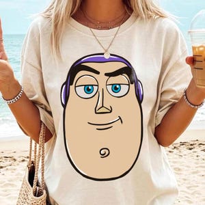 May include: Beige t-shirt featuring a large cartoon graphic of Buzz Lightyear's face. The graphic includes blue eyes, a purple headpiece, and a smiling expression. The shirt is a casual style.