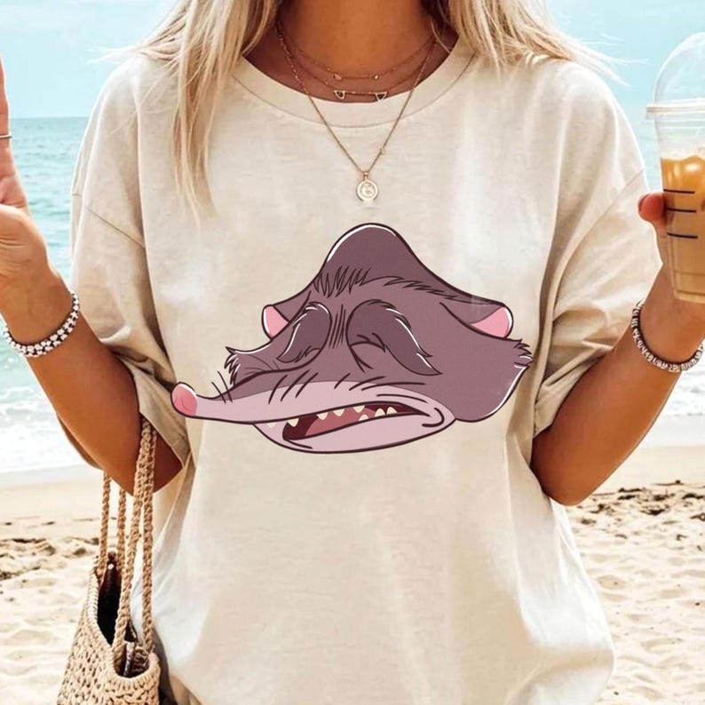 Peut inclure: T-shirt beige avec une illustration de dessin anim&eacute; d'un visage d'animal gris endormi avec des accents roses. Le dessin est centr&eacute; sur le devant du t-shirt. Le t-shirt est port&eacute; par une personne sur une plage.