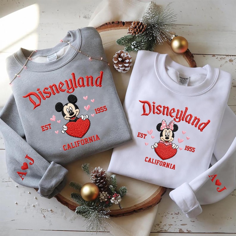 Puede incluir: Dos sudaderas, una gris y otra blanca, bordadas con "Disneyland" en escritura roja. Mickey Mouse y Minnie Mouse sostienen corazones rojos. El texto "EST 1955 CALIFORNIA" tambi&eacute;n est&aacute; bordado en rojo. La sudadera gris tiene las iniciales "A J" en la manga.