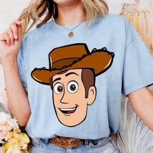 May include: Light blue t-shirt featuring a cartoon illustration of a cowboy character with a brown hat and smiling face. The graphic is centered on the shirt, creating a playful and fun design. The shirt is made of a soft material.