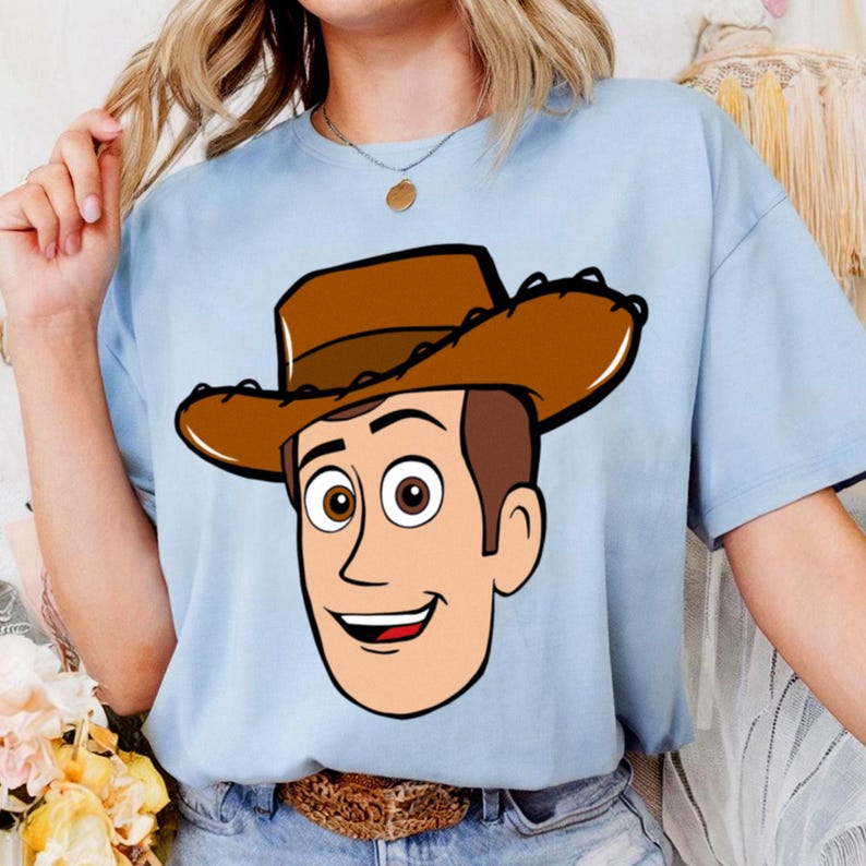May include: Light blue t-shirt featuring a cartoon illustration of a cowboy character with a brown hat and smiling face. The graphic is centered on the shirt, creating a playful and fun design. The shirt is made of a soft material.