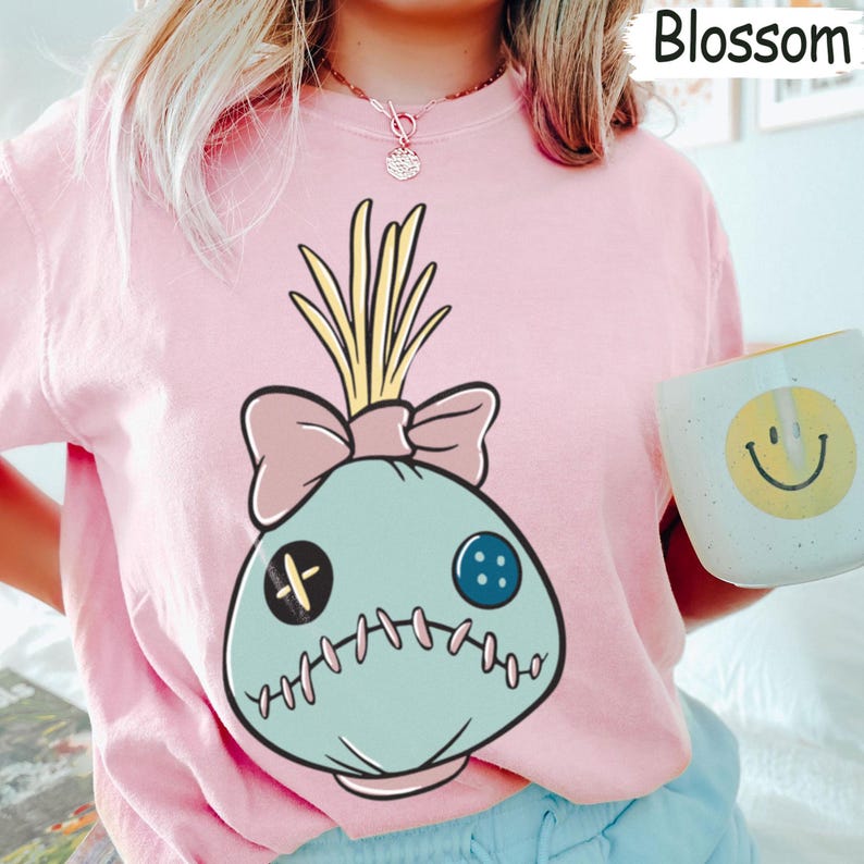 May include: A light pink sweatshirt featuring a cartoon character with a light blue head, a pink bow, and yellow hair. The character has a stitched mouth and button eyes. The word "Blossom" is printed in white on the top right.