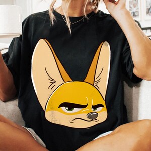 May include: Black t-shirt featuring a cartoon illustration of a fennec fox with large ears and a grumpy expression. The fox is primarily yellow and tan, with green eyes and a black nose. The shirt is a casual style.