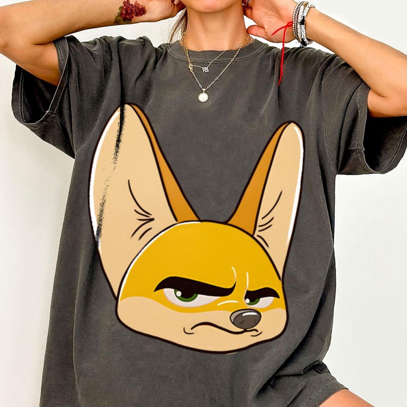 May include: Dark grey t-shirt featuring a cartoon fennec fox with large ears and a grumpy expression. The fox is primarily yellow and tan, with green eyes and a black nose. The shirt has short sleeves.