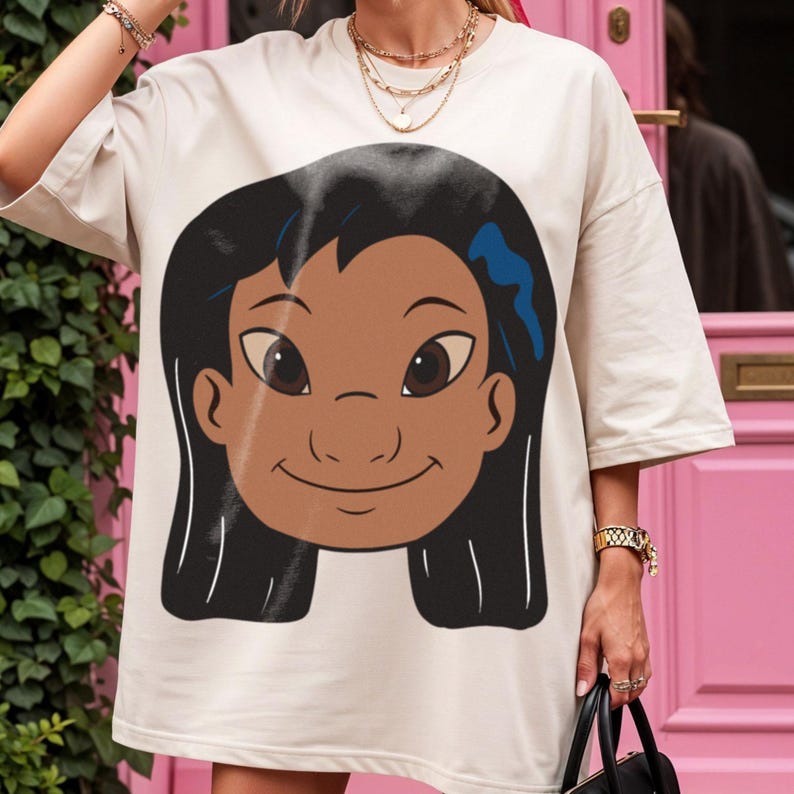 Puede incluir: Camiseta de color crema con una ilustraci&oacute;n de dibujos animados de una ni&ntilde;a sonriente con cabello negro, piel morena y ojos marrones grandes. El gr&aacute;fico est&aacute; centrado en la parte delantera de la camiseta. La camiseta tiene mangas cortas.