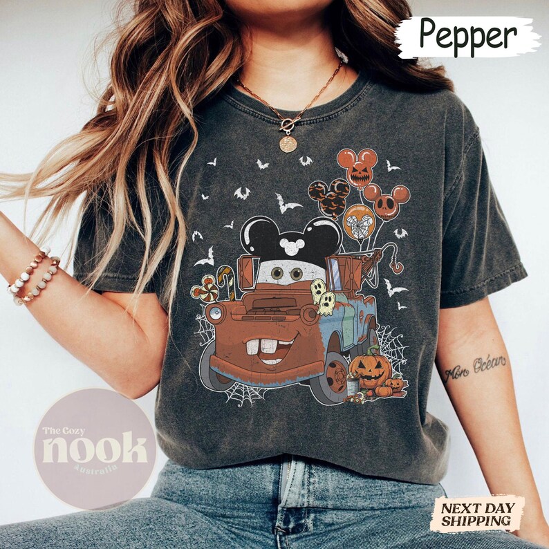 May include: Dark gray t-shirt featuring a cartoon image of Mater from the movie Cars, decorated for Halloween. The design includes Mickey Mouse-shaped balloons, bats, pumpkins, and ghosts. The text "Pepper" is at the top.