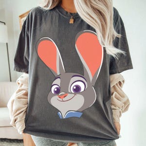 Puede incluir: Camiseta gris oscuro con un dibujo de la cara de un conejo de dibujos animados. El conejo tiene orejas grandes con el interior naranja, ojos morados y un cuello azul. La camiseta es de estilo casual.