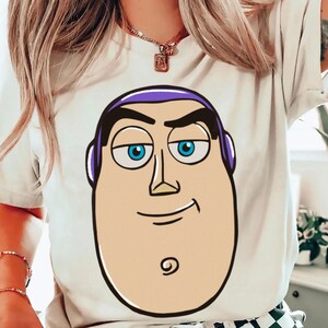 May include: White t-shirt featuring a large cartoon image of Buzz Lightyear's face. The graphic has a tan face, blue eyes, black eyebrows, and purple hair. The design is outlined in black.