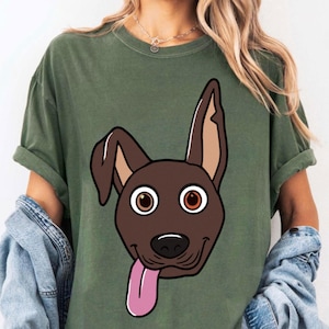 May include: Olive green t-shirt featuring a cartoon dog face with brown fur, large brown eyes, and a pink tongue sticking out. The dog's ears are a lighter brown color. The shirt has rolled-up sleeves and is worn with a denim jacket.