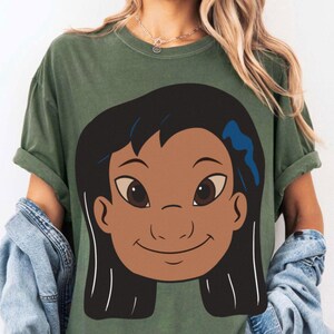 Puede incluir: Camiseta verde oliva con una ilustraci&oacute;n de dibujos animados de una ni&ntilde;a sonriente con piel morena, cabello negro y un accesorio azul para el cabello. El gr&aacute;fico est&aacute; centrado en la parte delantera. La camiseta tiene mangas cortas y un corte holgado.